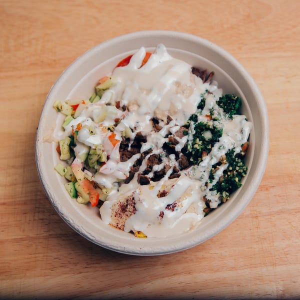 A Jarasa Kabob bowl with meat, tabbouleh, chopped salad, and white sauce on a wooden table.